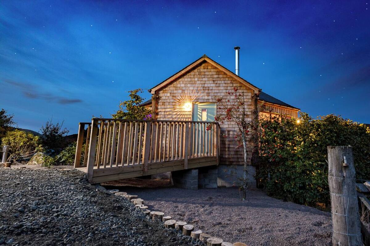 A rustic cabin with a wooden deck, a chimney, and a gravel driveway.