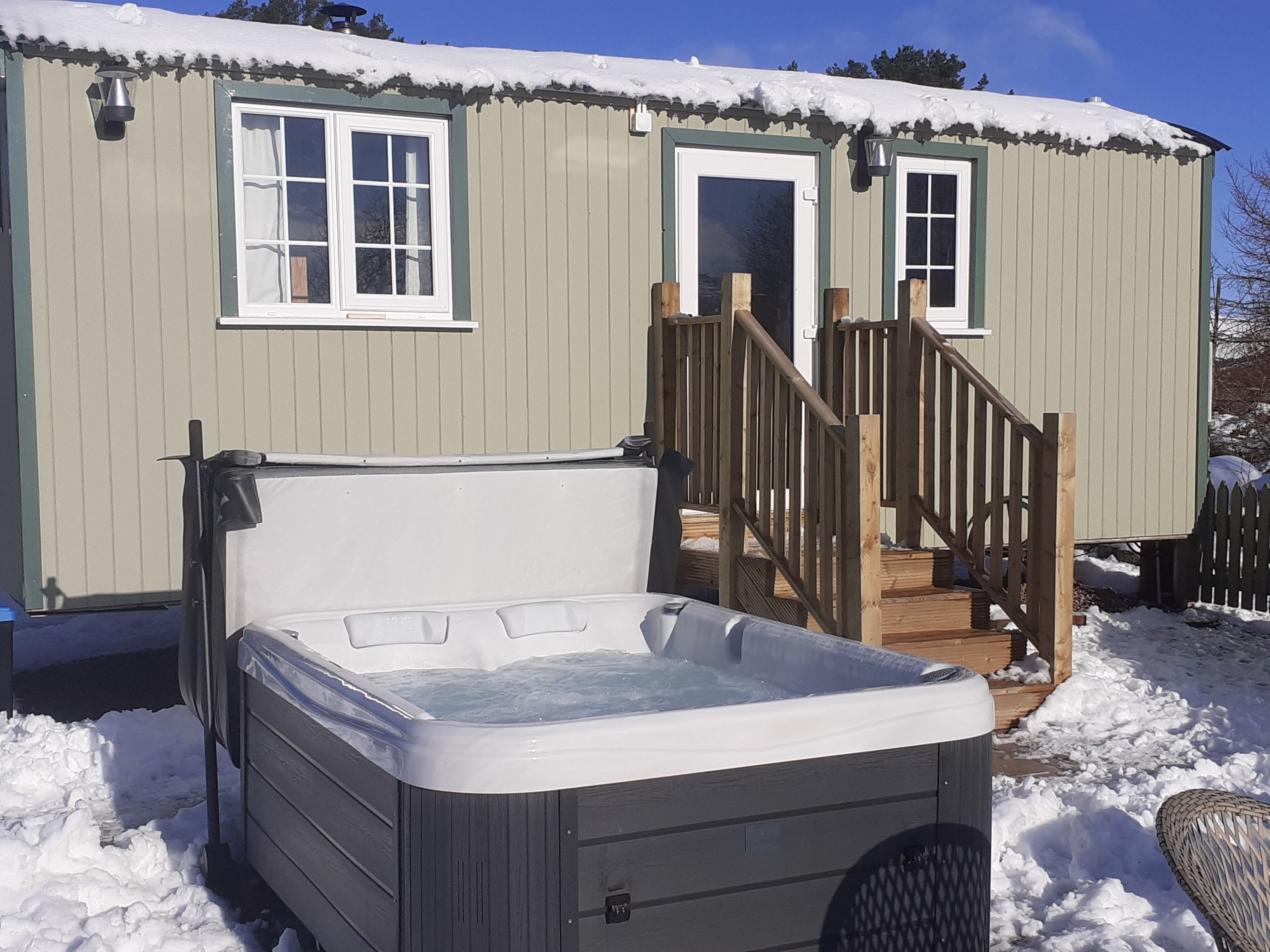 A hot tub in front of a wooden cabin with a deck and a staircase leading to the door.