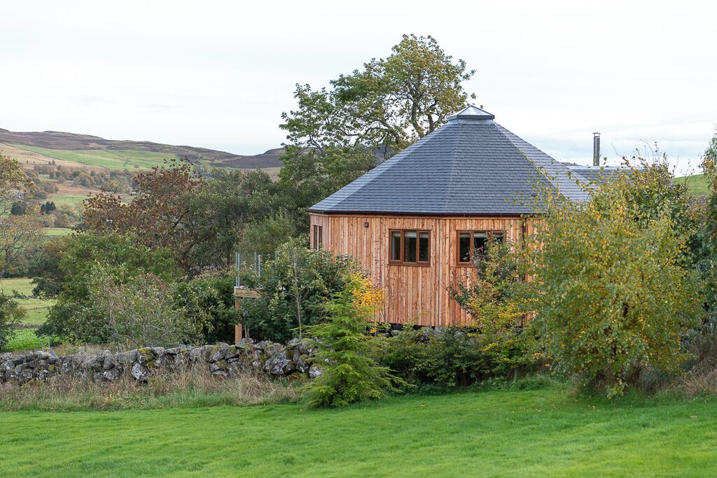 A wooden cabin with a sloped roof, surrounded by a stone wall and lush greenery.