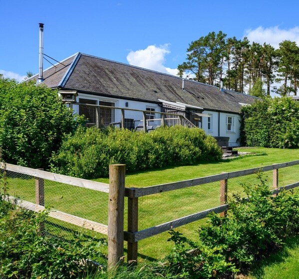 A two-story house with a sloped roof, surrounded by lush greenery and a wooden fence. - Dalnoid Cottages & Treehouses (Blairgowrie)