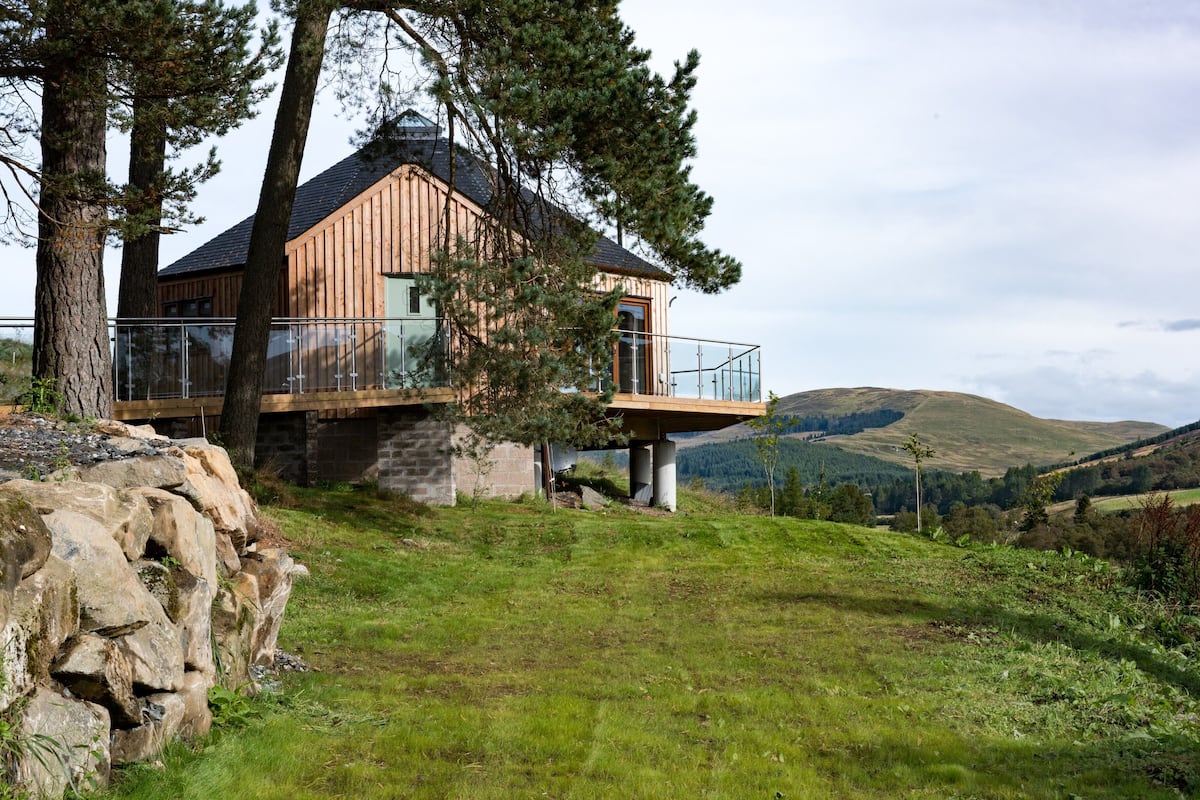 A wooden cabin with a balcony, surrounded by trees and hills.
