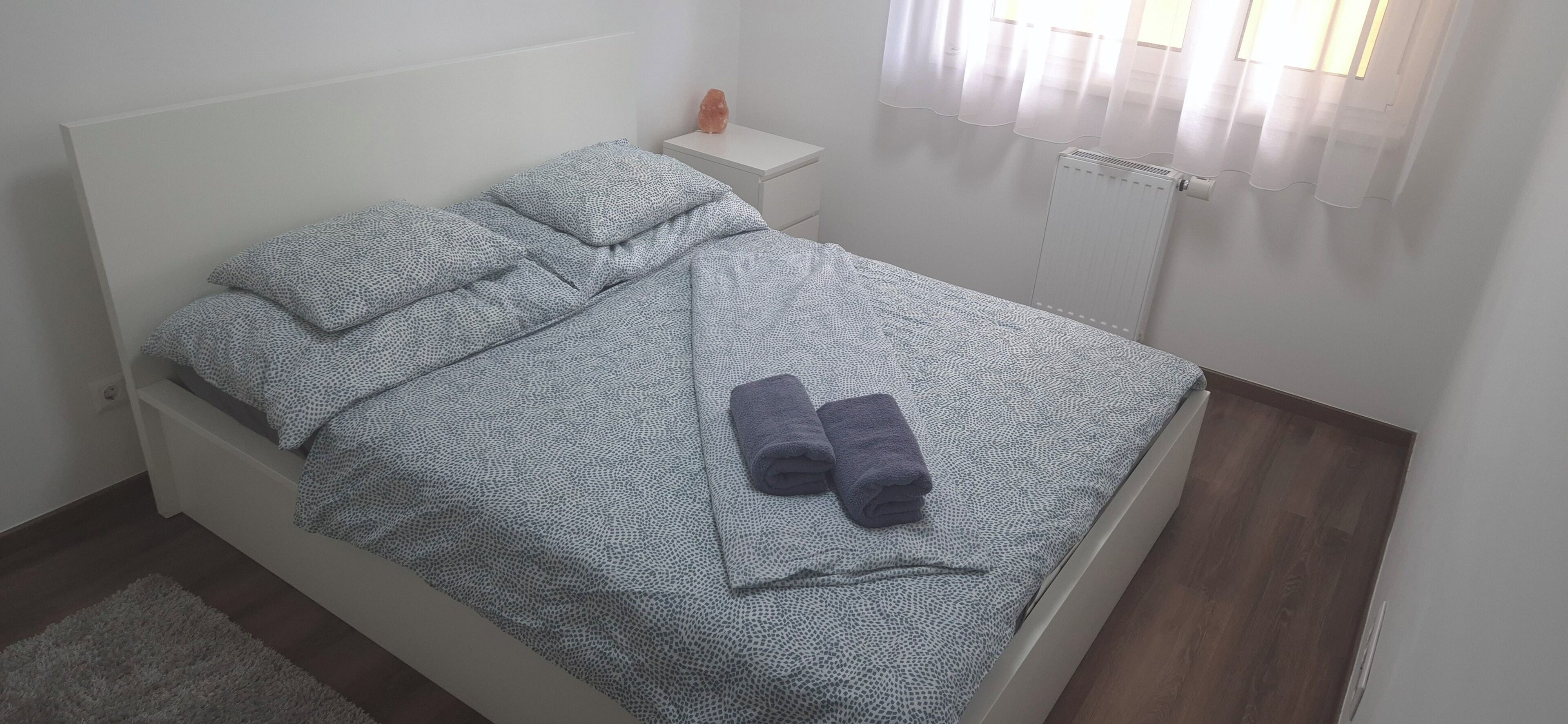 A neatly made bed with patterned bedding and rolled towels, a white bedside table with a small item, and a radiator in the corner.