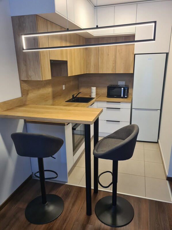 A modern kitchen with wooden cabinets, a black countertop, a microwave, and a refrigerator. There are two bar stools and a sink with a faucet. - Aphrodite Apartman Budapest (Budapest)