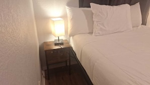 Una cama bien hecha con ropa blanca, una mesita de noche de madera con una lámpara y un suelo de madera oscura.