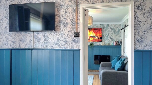 A room with a blue wall, a flat-screen TV mounted above a floral-patterned wallpaper, a grey sofa with blue cushions, and a fireplace with a fire.