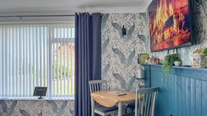 A dining area with a wooden table and chairs, a window with blinds, a wall with a peacock pattern, and a mounted TV displaying a fire scene.