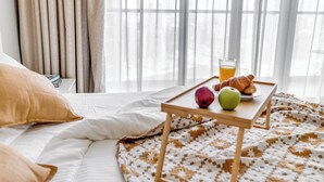 A neatly made bed with a white comforter and a patterned blanket, a bedside table with breakfast items including croissants, an apple, and a glass of orange juice, and a window with sheer curtains allowing natural light.