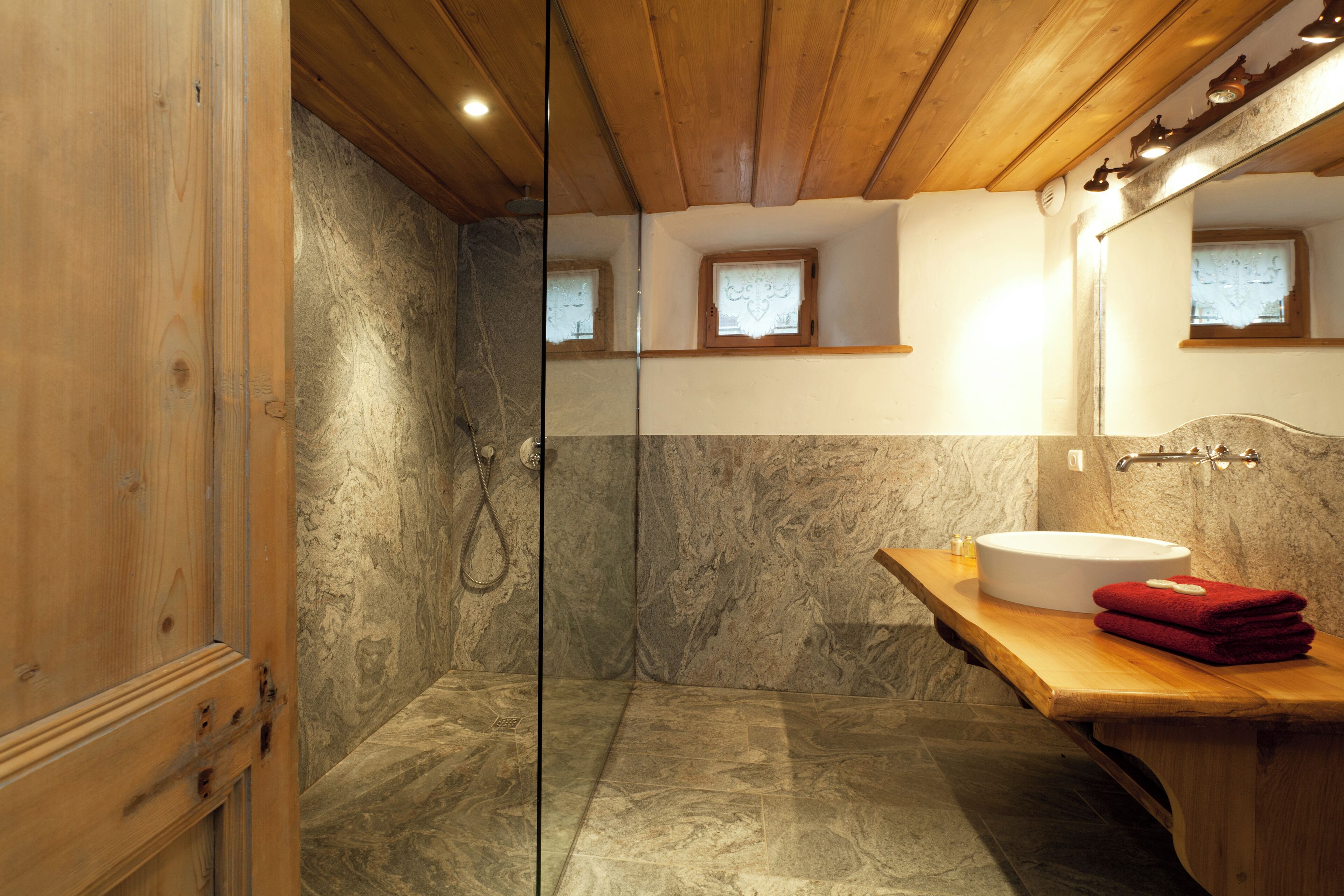 A bathroom with a wooden ceiling, a glass shower enclosure, a marble wall, a wooden vanity, and a mirror.
