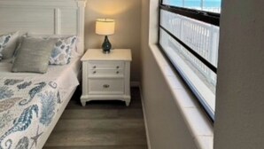 Un dormitorio con cama, mesita de noche, lámpara y ventana con vista a árboles.