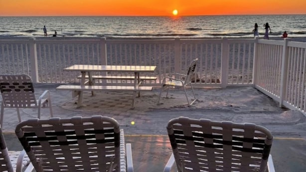 Un patio frente a la playa con una mesa y sillas, con vista al océano al atardecer.