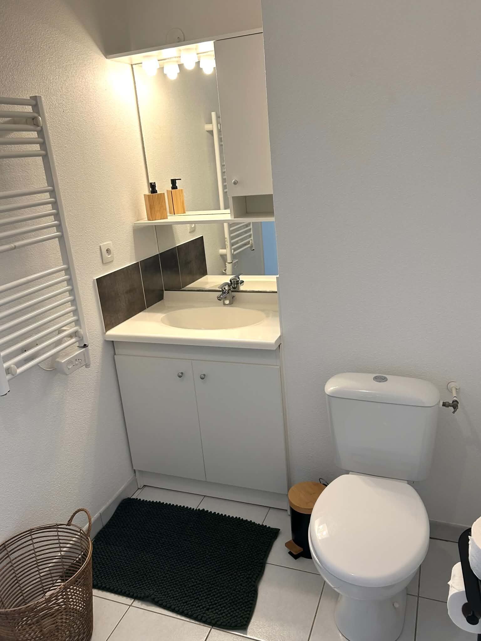 Ein Badezimmer mit einem weißen Waschbecken, einem Spiegel, einer Toilette und einer schwarzen Badmatte.