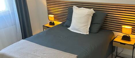 Una cama bien tendida con un edredón de color oscuro, dos almohadas y una manta blanca. Hay dos mesitas de noche con lámparas, y el suelo está cubierto de baldosas.