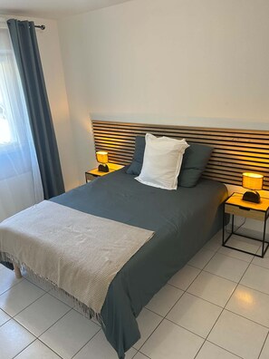 A neatly made bed with a dark-colored duvet, two pillows, and a white throw blanket. There are two bedside tables with lamps, and the room has a tiled floor.