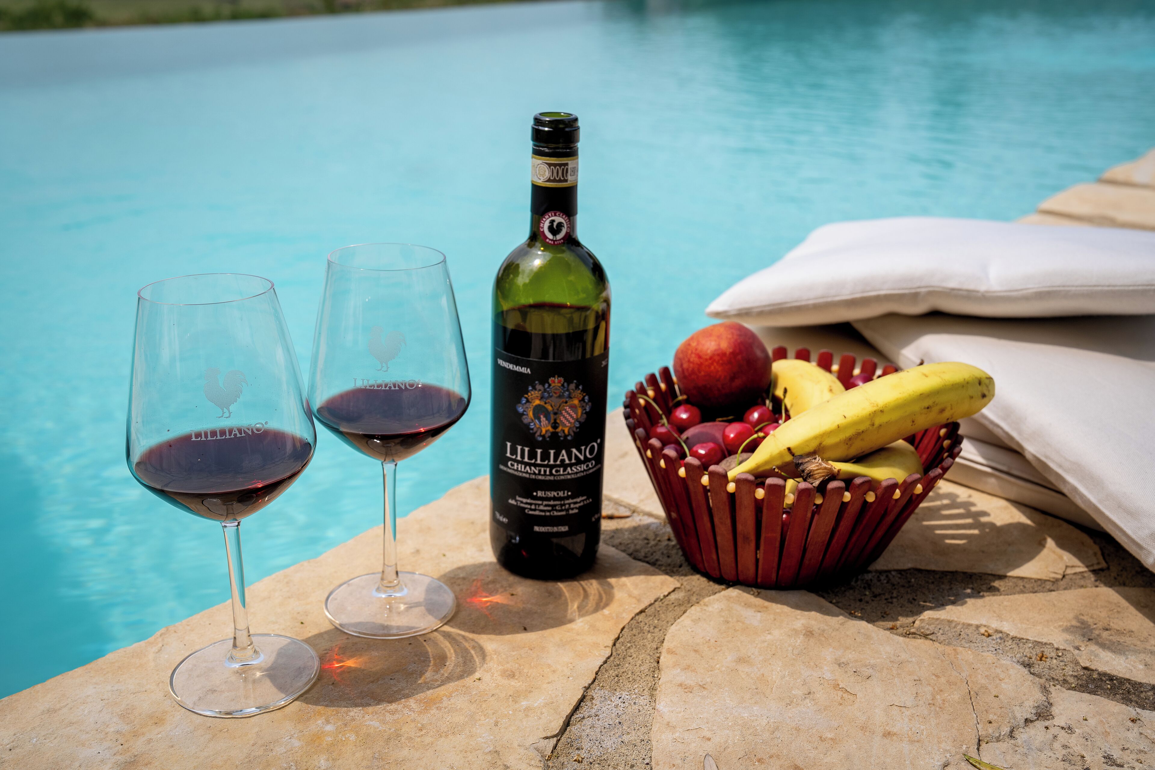 Zwei Weingläser mit Rotwein, eine Flasche Lilliano Chianti Classico Wein und ein Obstkorb mit Bananen und Äpfeln, am Pool gelegen.