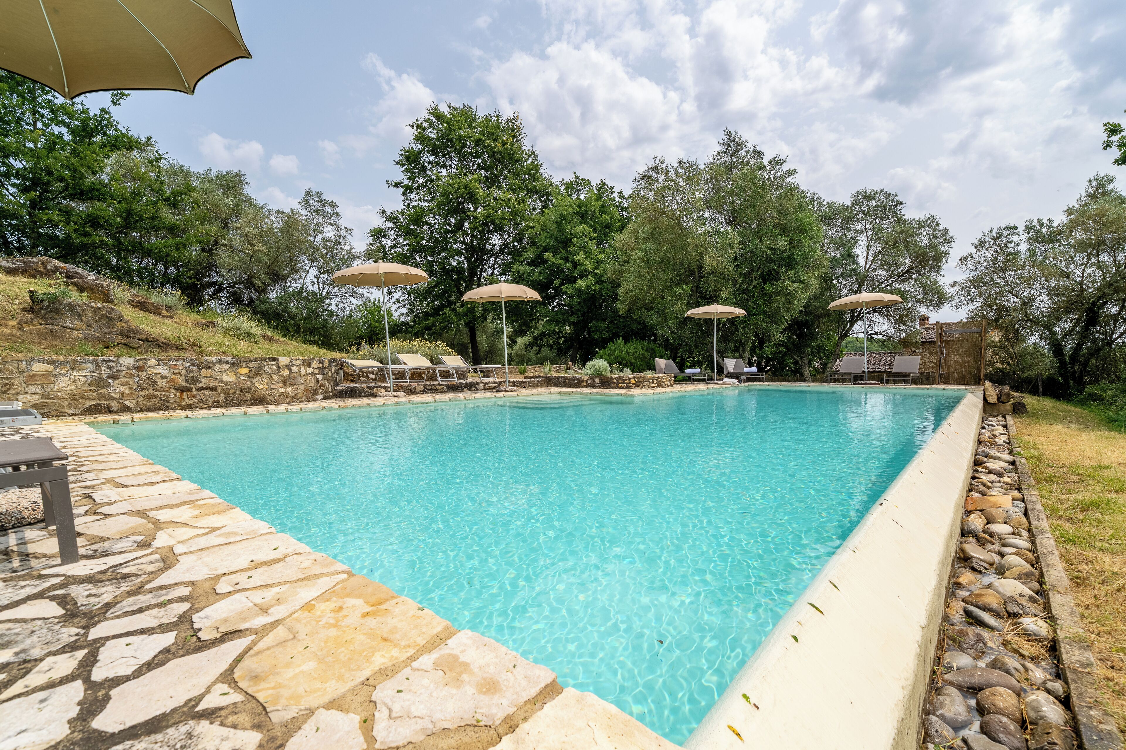 A clear blue pool with lounge chairs and umbrellas, surrounded by stone walls and trees.