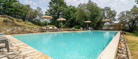 A clear blue pool with lounge chairs and umbrellas, surrounded by stone walls and trees.