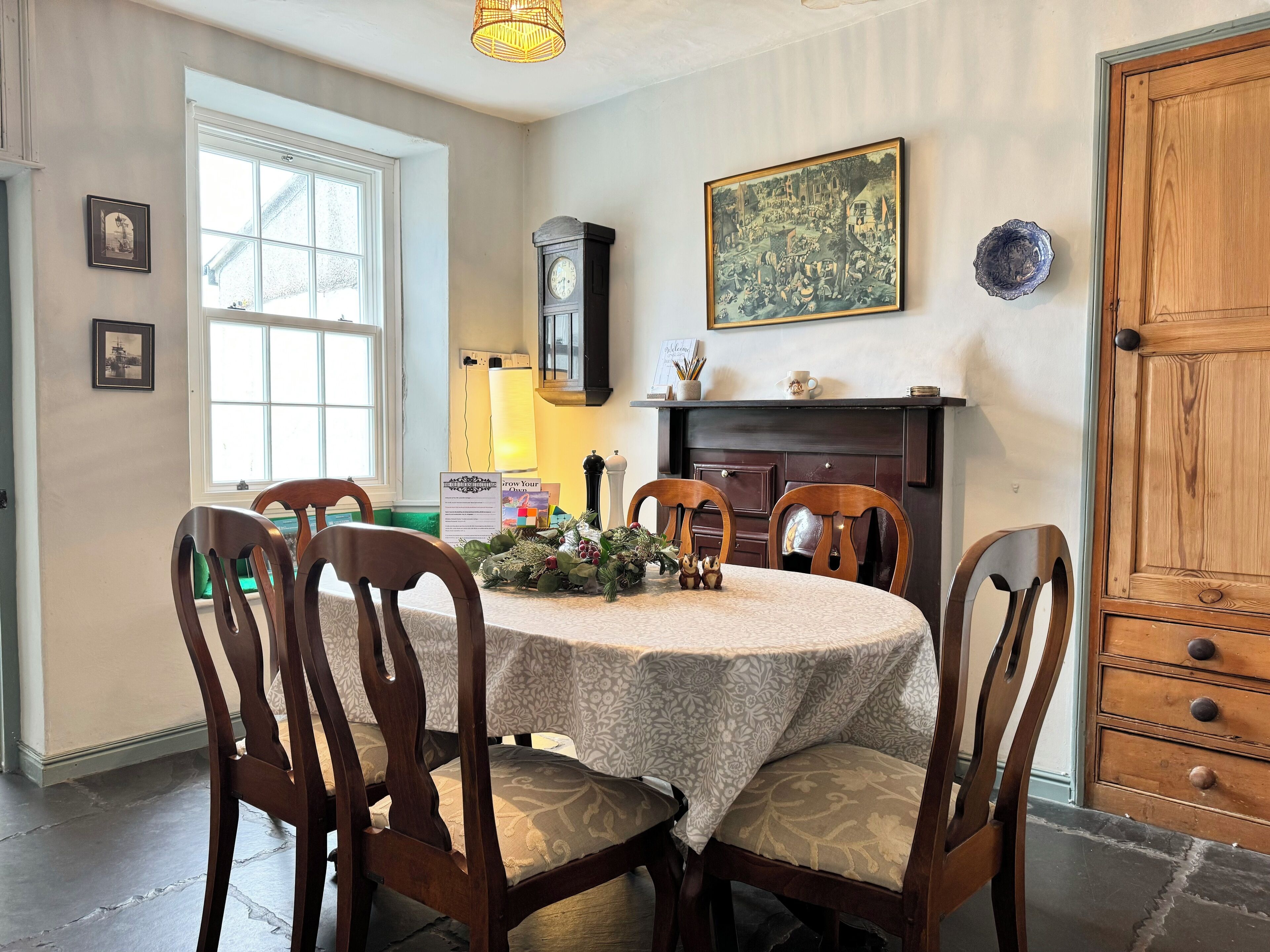 A dining room with a round table, wooden chairs, a clock, a fireplace, and framed artwork on the wall.