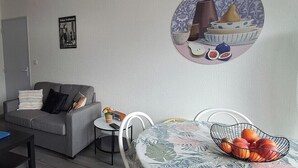 A living room with a grey sofa, a small round table with a white chair, a black coffee table with a blue object, and a fruit basket on a table with a floral tablecloth.