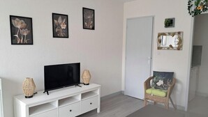 A living room with a flat-screen TV on a white TV stand, a wicker chair with a green cushion, and three framed floral artworks on the wall.