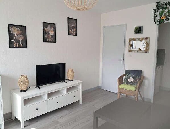 Una sala de estar con un televisor de pantalla plana sobre un mueble blanco, un sillón de mimbre con un cojín verde, y tres cuadros con motivos florales en la pared.