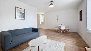 A modern living room with a blue sofa, a small white table, and a white chair. There is a large window with a view of the outside. - Bright 1-bedroom apartment (Cuxac-d'Aude)