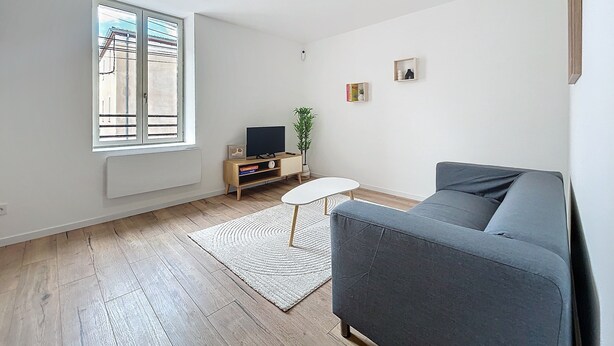 A living room with a grey sofa, a small white table, a TV on a wooden stand, and a window with a view of buildings. - Bright 1-bedroom apartment (Cuxac-d'Aude)