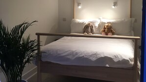Een bed met houten frame, witte beddengoed en twee knuffeldieren. Links van het bed staat een pot met een plant.