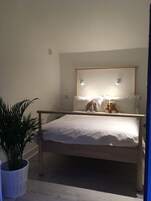 A bed with a wooden frame, white bedding, and two stuffed animals. There is a potted plant to the left of the bed.