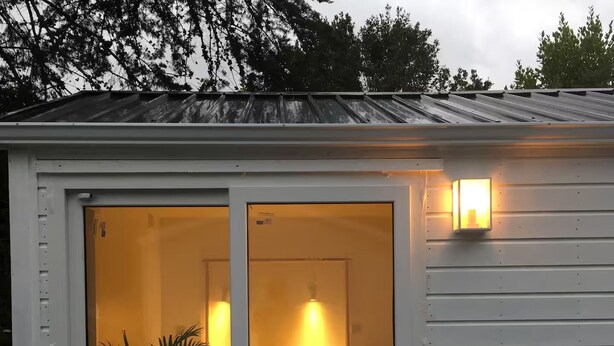 Een klein, vrijstaand vakantiehuisje met een schuifpui, een bed, een nachtkastje en een bloempot.