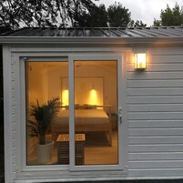 A small, single-room cabin with a sliding glass door, a bed, a bedside table, and a potted plant.