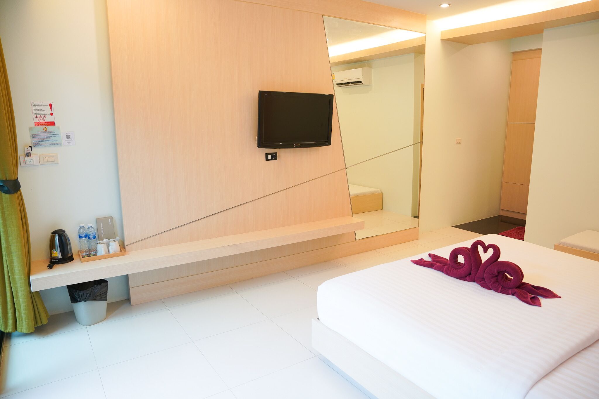 A hotel room with a bed, a flat-screen TV mounted on the wall, a small table with a kettle and bottles, and a trash bin.