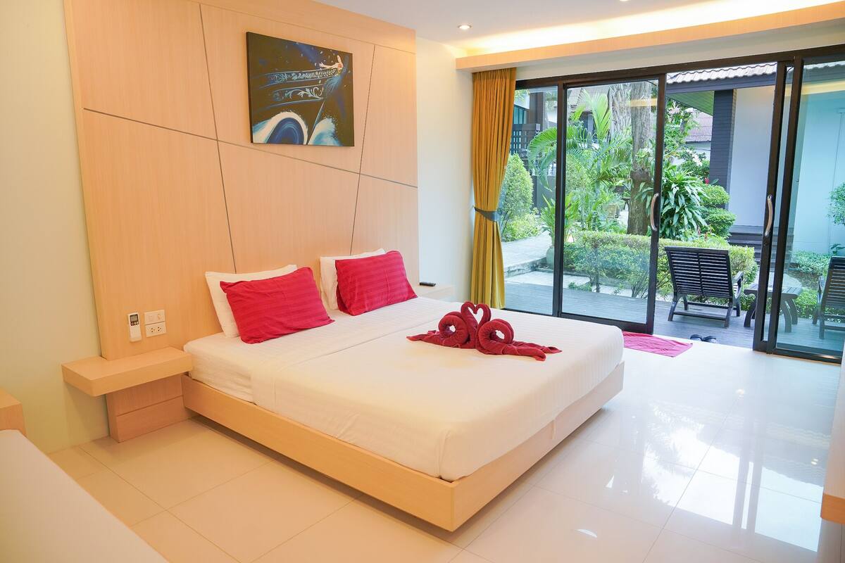 Una habitación de hotel moderna con una cama grande, una mesita de noche, un cuadro en la pared y vistas al jardín a través de la puerta de vidrio.