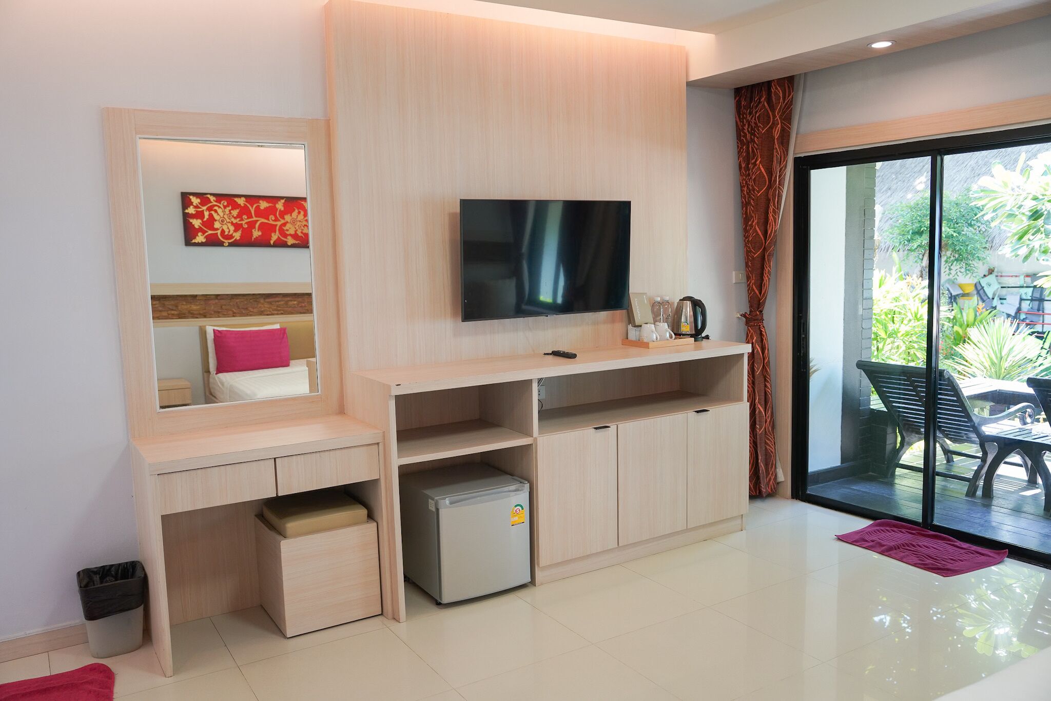 A modern hotel room with a flat-screen TV, a small refrigerator, a desk with a chair, and a large sliding glass door leading to an outdoor area with a table and chairs.
