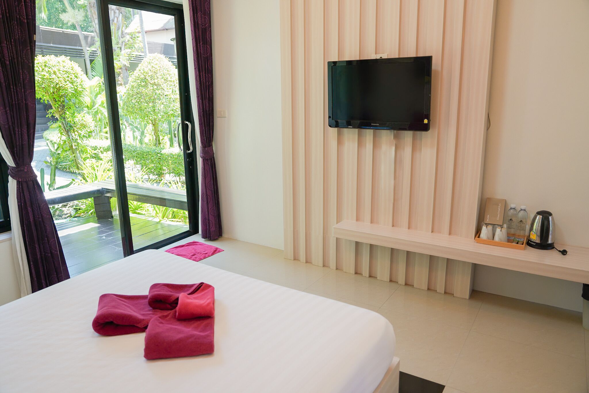A hotel room with a bed, a TV mounted on the wall, a bench with toiletries, and a view of greenery outside.
