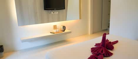 Una habitación de hotel moderna con un televisor de pantalla plana montado en la pared, una mesita de noche con un hervidor, y una cama bien hecha con una toalla roja.