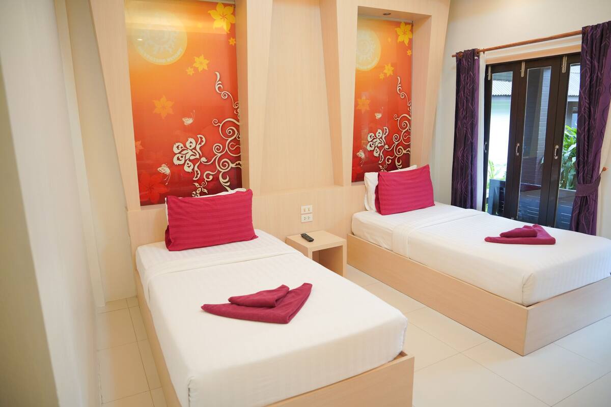 Habitación de hotel con dos camas individuales, cada una con colchón blanco y almohada roja. Se observa una pequeña mesita de noche con un control remoto. La habitación tiene un ventanal grande con cortinas moradas y vistas al exterior.