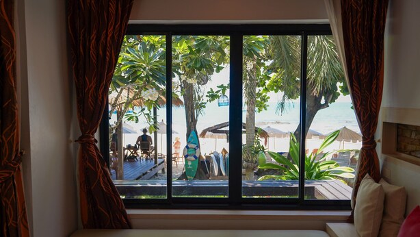 Ein Zimmer mit großem Fenster und Blick auf einen Strand, einer gepolsterten Bank sowie Strandsonnenschirme und Palmen.