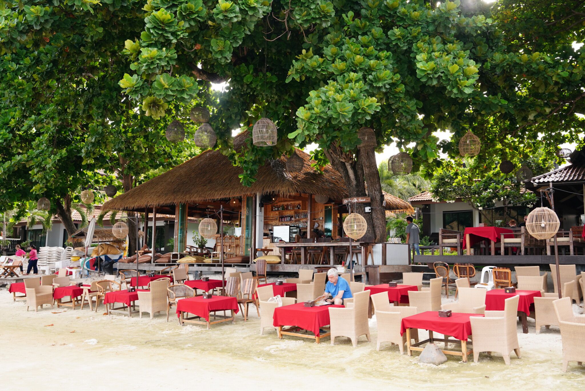 An outdoor restaurant with tables and chairs, a thatched-roof building, and hanging birdcages.