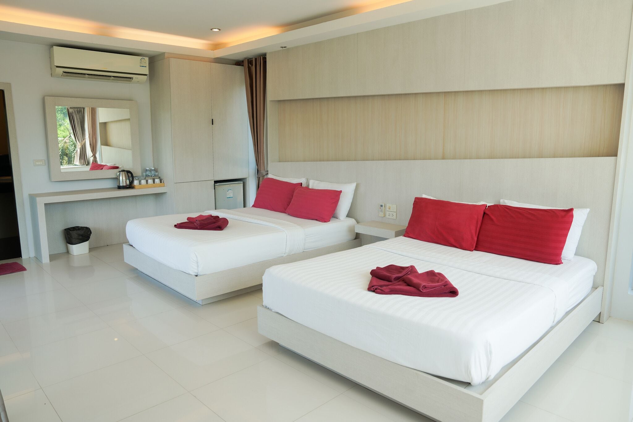 A hotel room with two beds, each with red pillows, a large mirror, a small table with a kettle, and a trash bin.