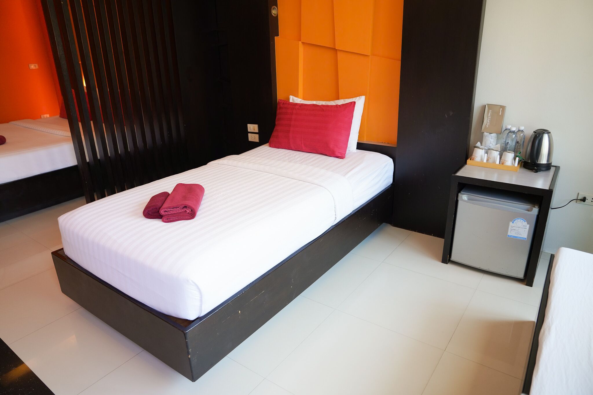 A hotel room with a bed, a small bedside table with a kettle, and a mini fridge.