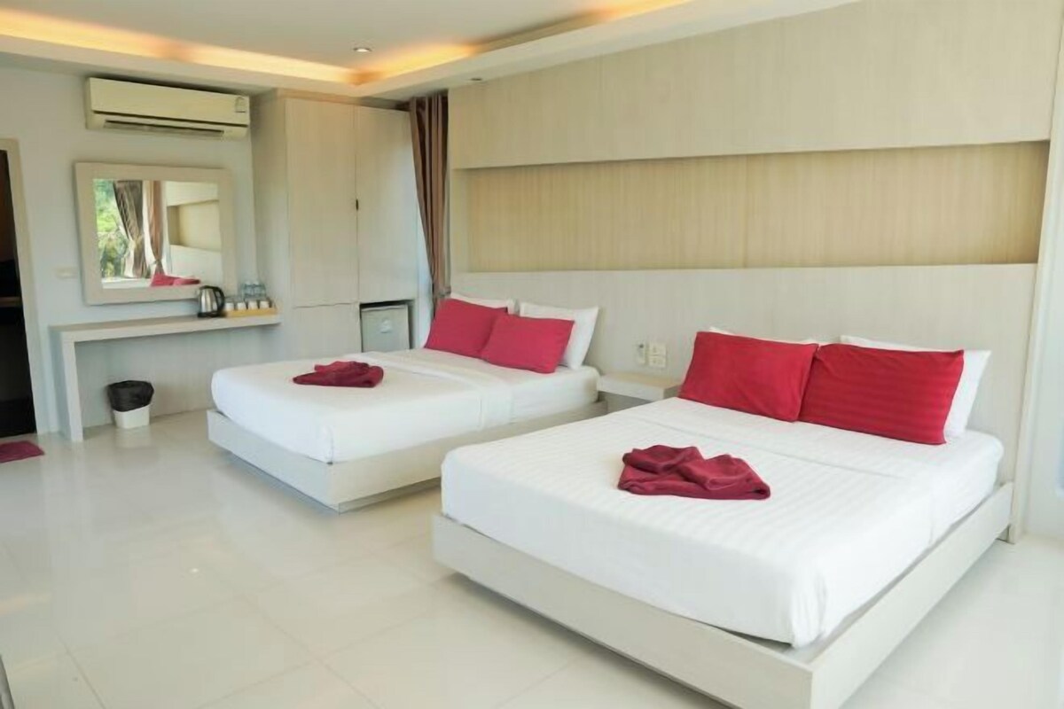 A hotel room with two beds, each with white bedding and red pillows. There is a mirror above the beds, a small table with a lamp, and a trash bin.