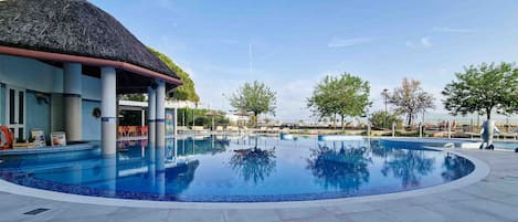A swimming pool with a thatched roof structure, lounge chairs, and a clear sky.