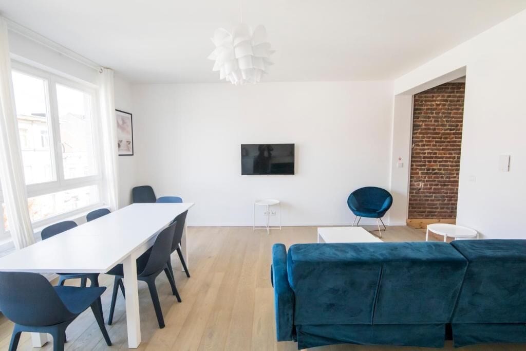 Una sala de estar moderna con un sofá blanco, una mesa de comedor con sillas, un televisor montado en la pared y una pared de ladrillo.
