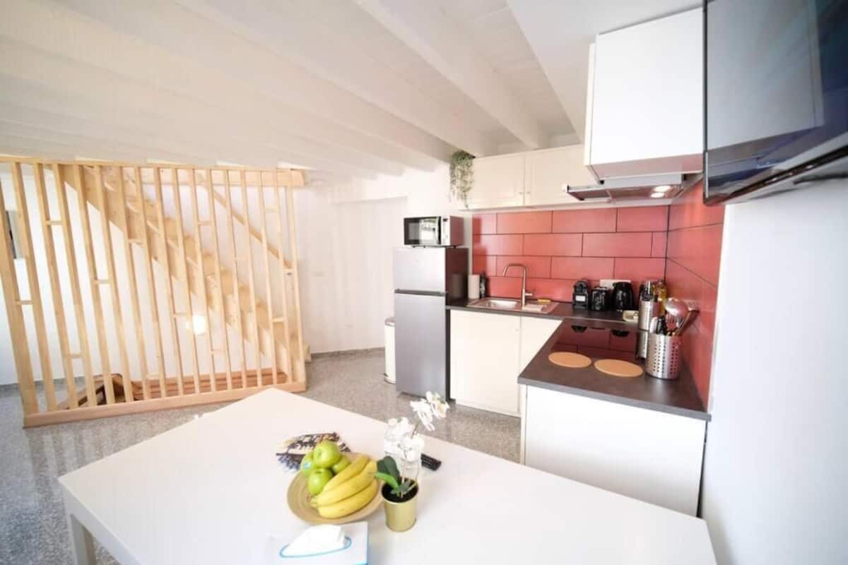 A modern kitchen with white cabinets, a red backsplash, and a wooden dining table with fruit and a vase.