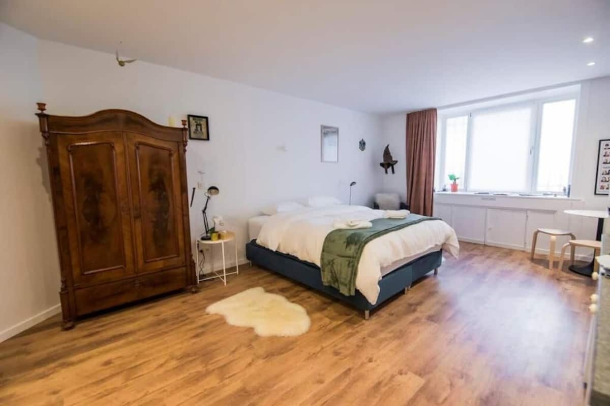 A bedroom with a wooden wardrobe, a bed with a white and green blanket, a small bedside table with a lamp, and a window with curtains.
