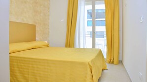 A room with a bed, a window with yellow curtains, and a view of a building outside. - Salt air and Soft Light Flat in Sunny Bibione (San Michele al Tagliamento)