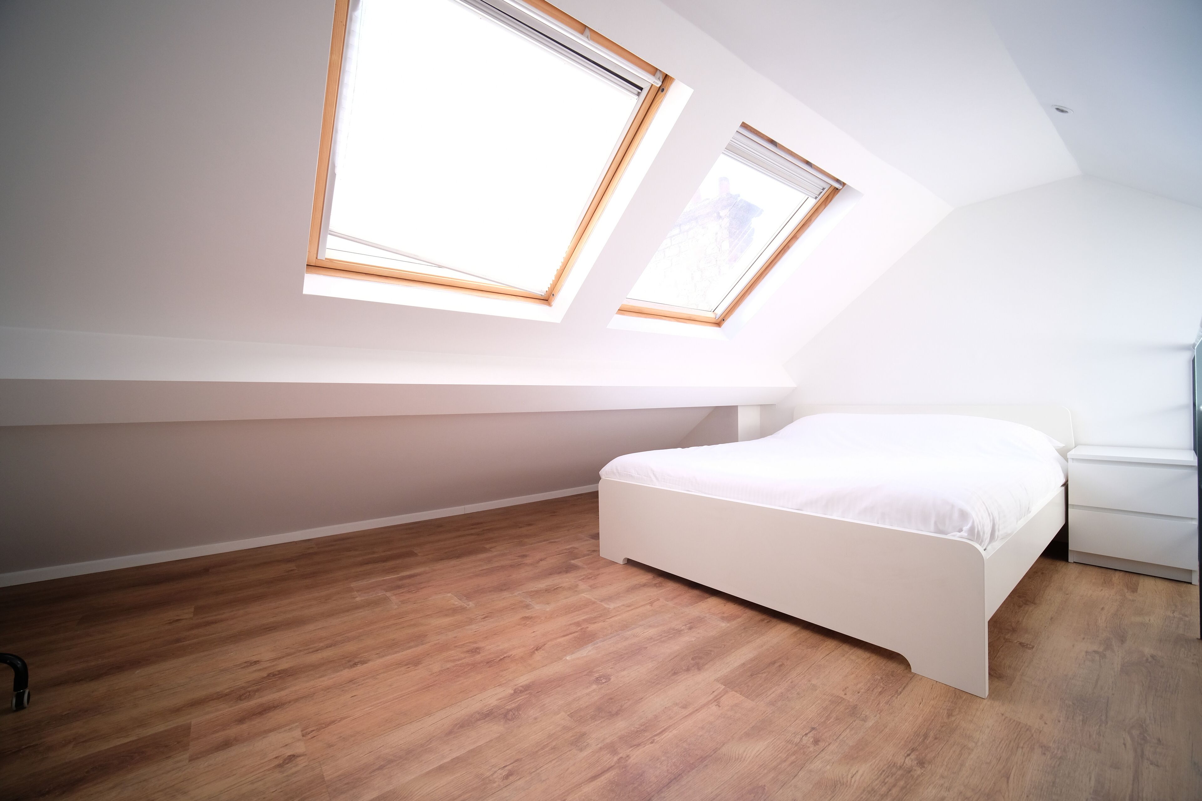 Une chambre avec un plafond en pente, deux fenêtres de toit, un lit blanc et un plancher en bois.