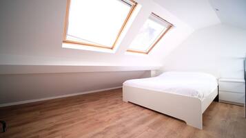 Une chambre avec un plafond en pente, deux fenêtres de toit, un lit blanc et un plancher en bois.