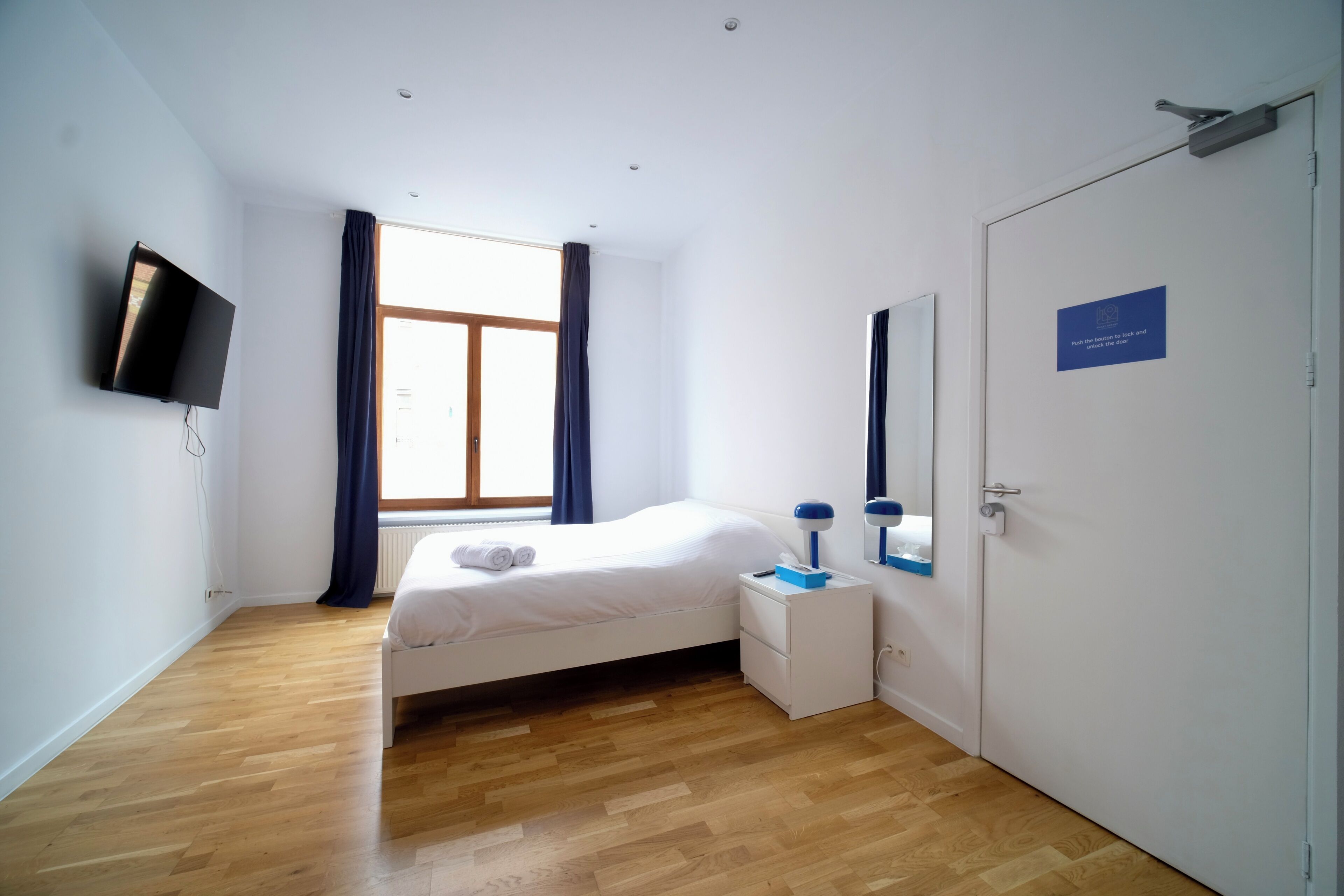 Een slaapkamer met een bed, nachtkastje, lamp, televisie, raam met gordijnen en een deur met een bordje.