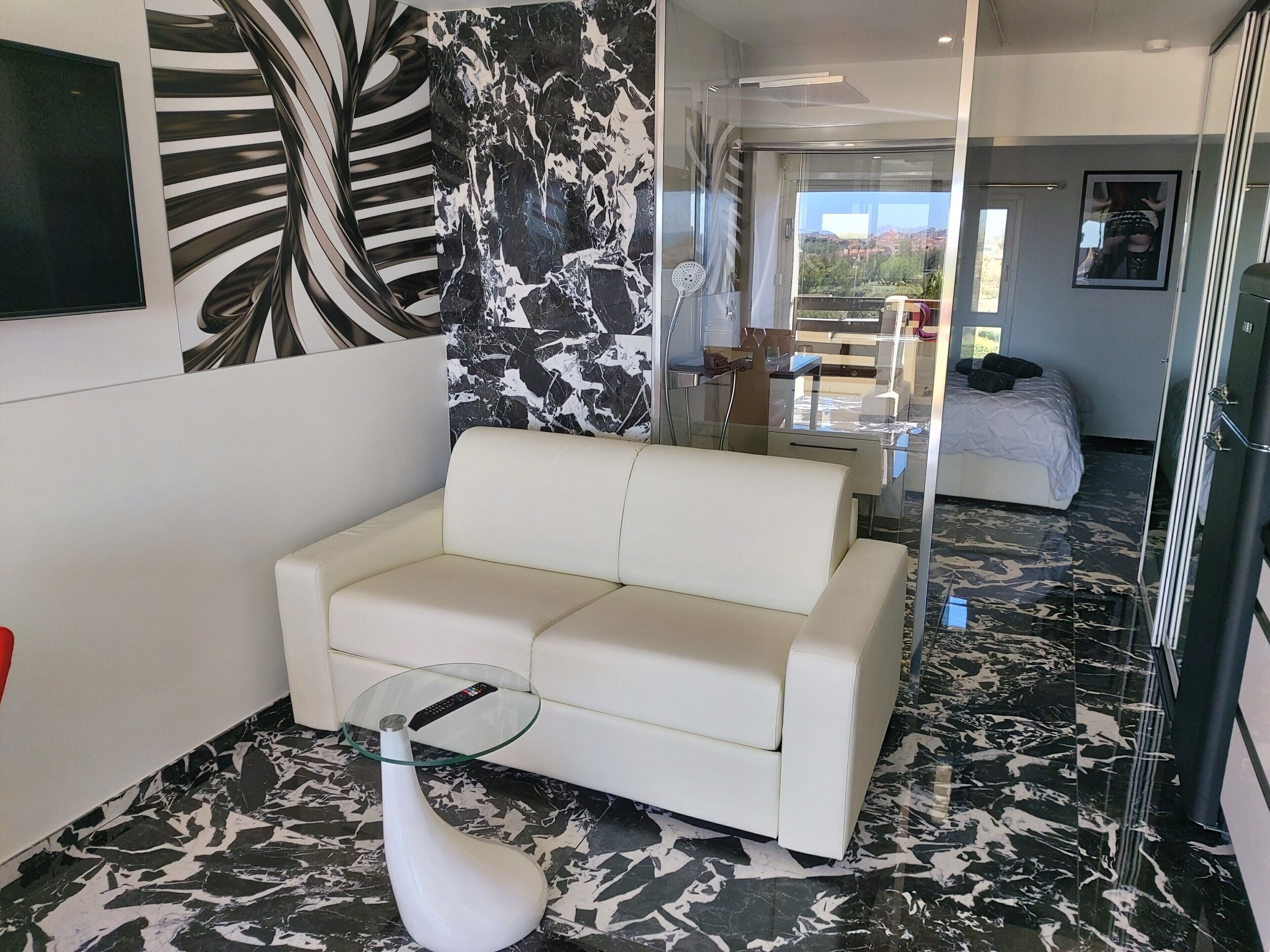 A modern hotel room with a white sofa, a glass coffee table, a white vase, and a large mirror reflecting the room. The floor has a black and white marble pattern, and there is a bed with a white duvet in the background.